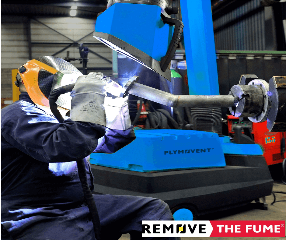 A man welding with a protective mask on and in the background there is a blue Remove The Fume Fume Extractor who's extraction arm is sucking all the harmful fumes away.