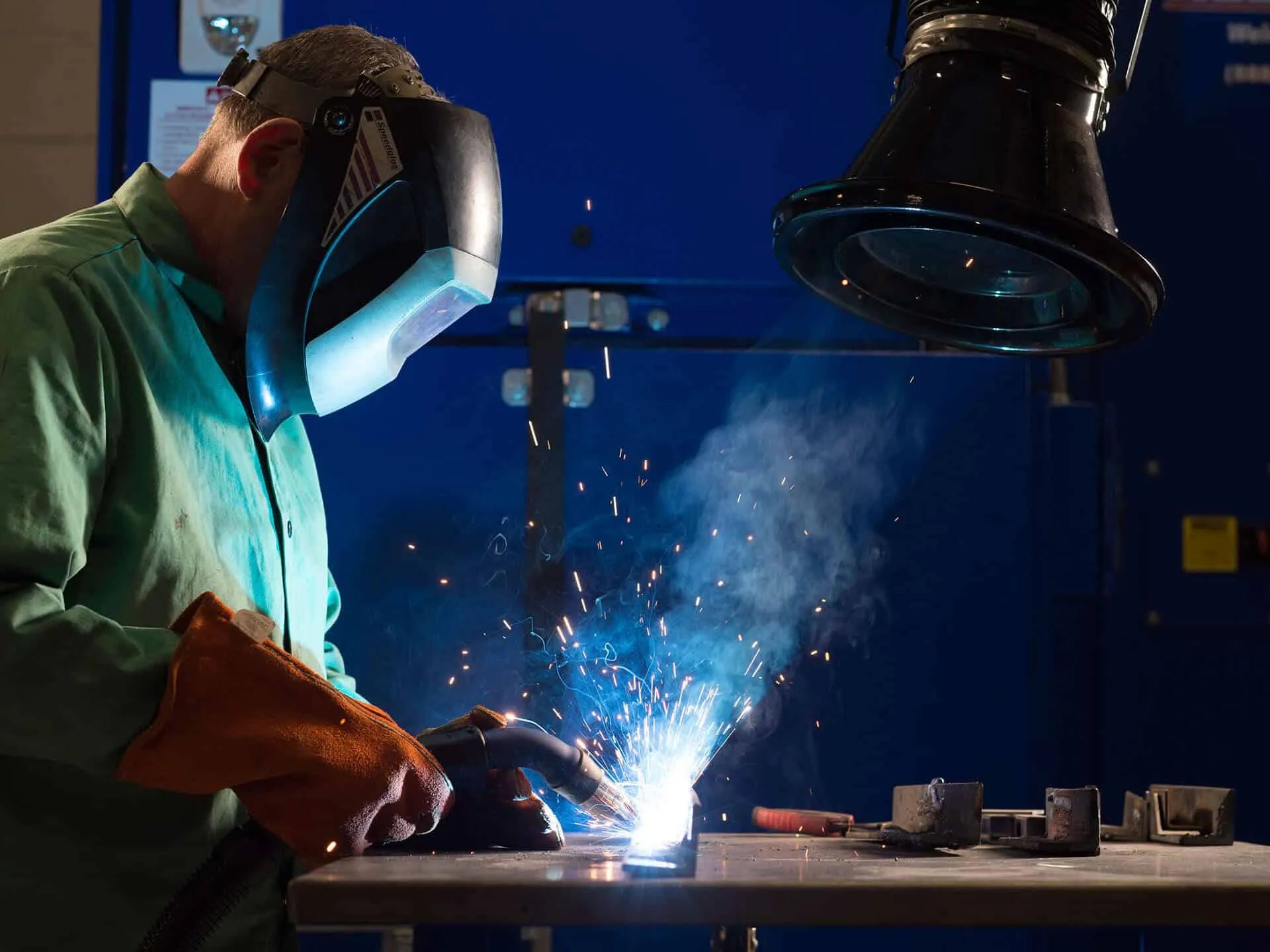 A man welding with a protective mask on and there are welding fume exerted which are being captured by the black colored fume extraction arm.
