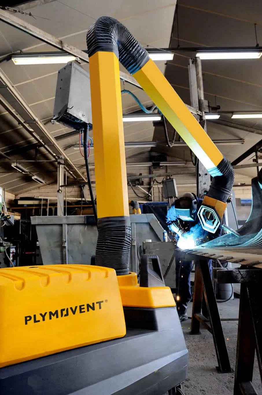 A man welding with a protective mask on a while yellow Remove The Fume fume extractor arm is inhaling all the harmful fume away.
