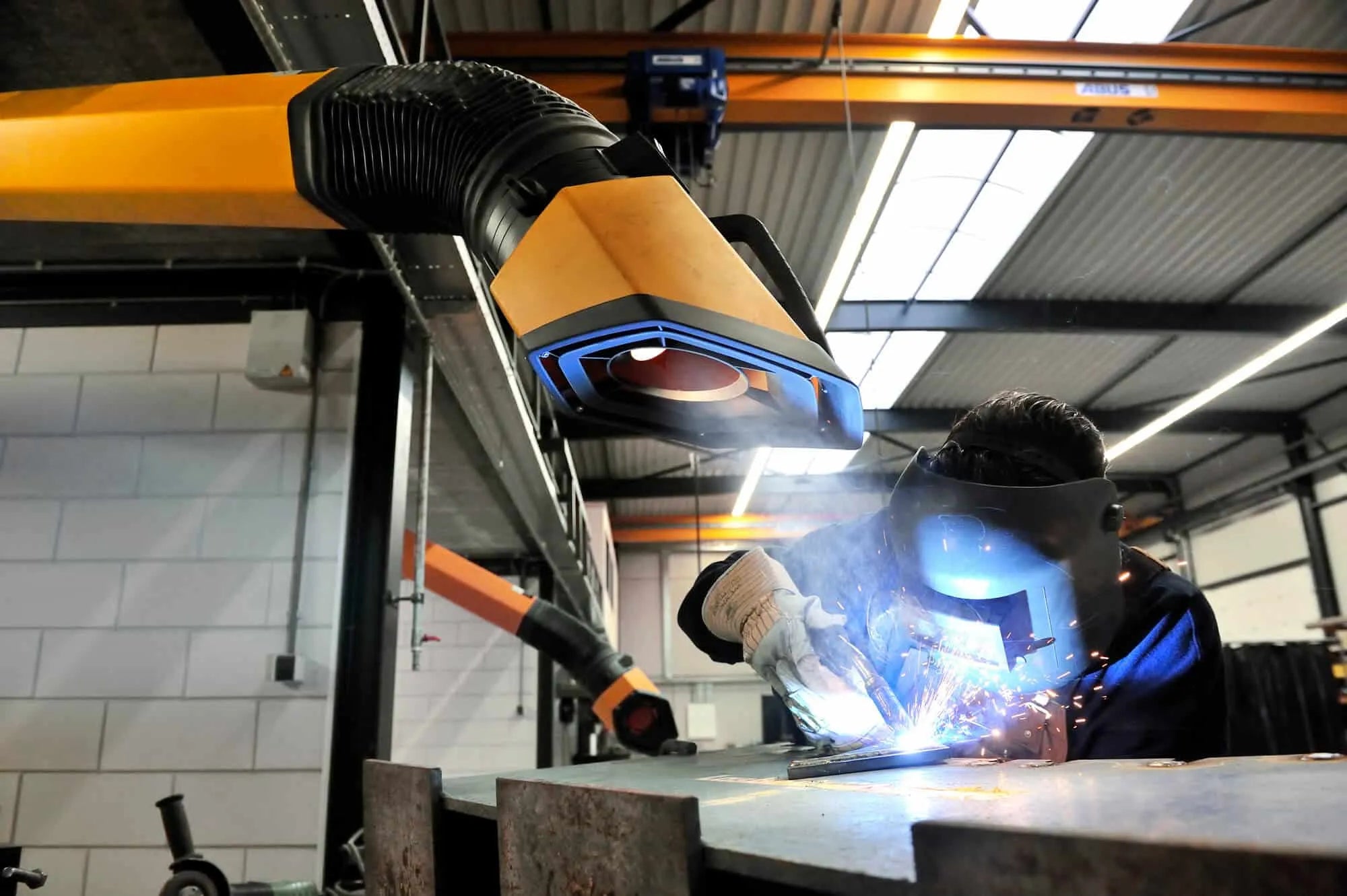 A man welding with a protective mask on a while yellow Remove The Fume fume extractor arm is inhaling all the harmful fume away.