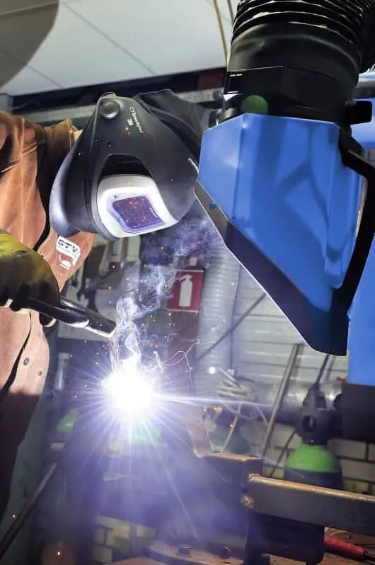 A man welding with a protective mask on and in the background there is a blue Remove The Fume Fume Extractor who's extraction arm is sucking all the harmful fumes away.