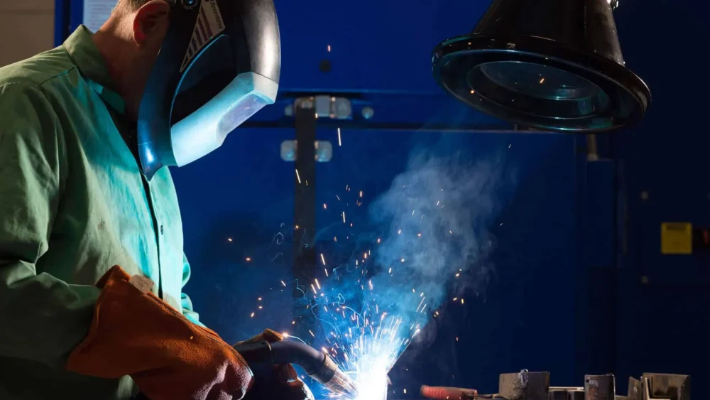 A man welding with a protective mask on and there are welding fume exerted which are being captured by the black colored fume extraction arm.