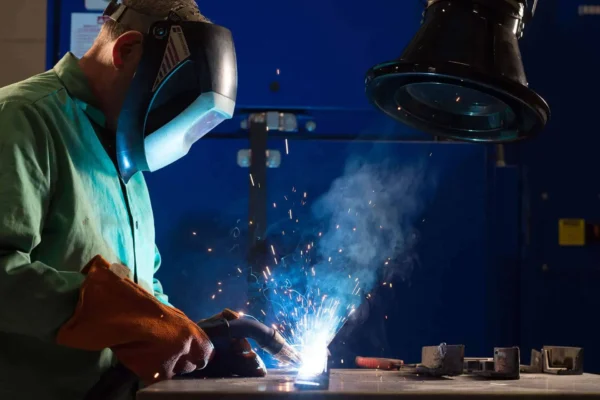 A man welding with a protective mask on and there are welding fume exerted which are being captured by the black colored fume extraction arm.