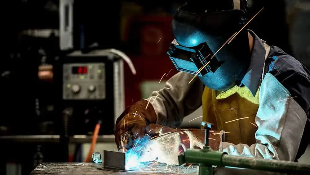 A man welding with a black protective mask on while ther eare harmful sparks and fumes being generated.