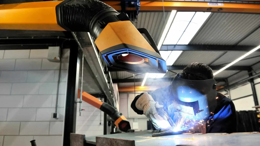 A man welding with a protective mask on a while yellow Remove The Fume fume extractor arm is inhaling all the harmful fume away.