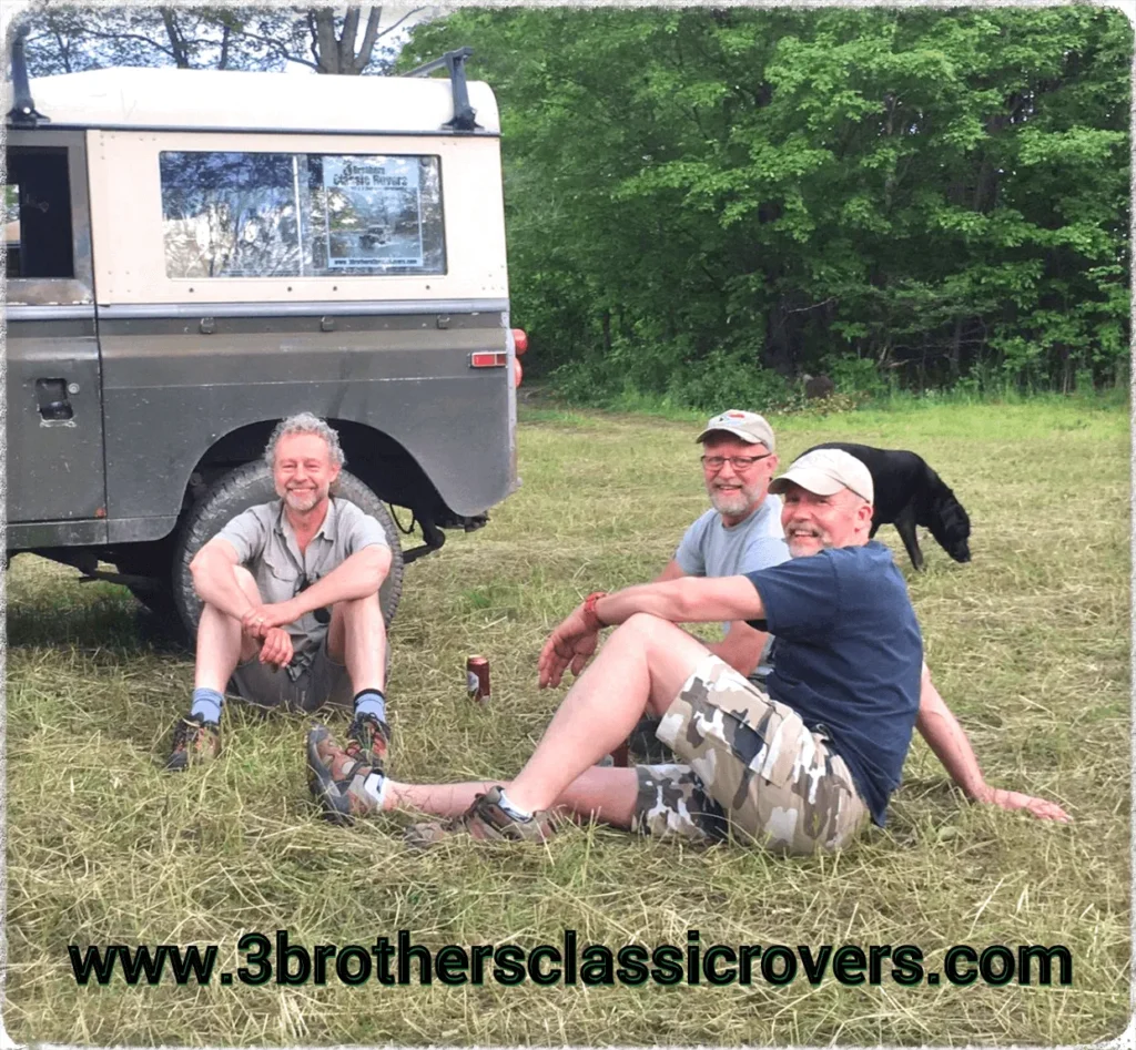 three men sitting in an open field next to a car with a black colored dog in the background.