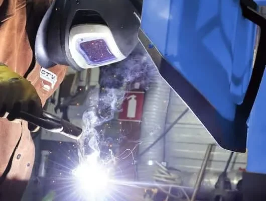 A man welding with a protective mask on and in the background there is a blue Remove The Fume Fume Extractor who's extraction arm is sucking all the harmful fumes away.