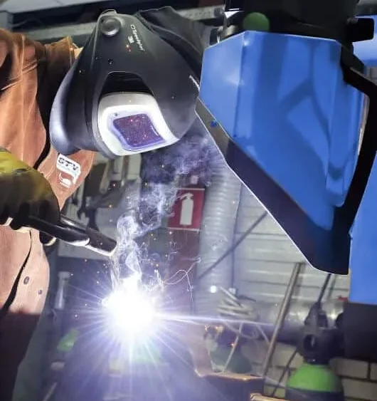 A man welding with a protective mask on and in the background there is a blue Remove The Fume Fume Extractor who's extraction arm is sucking all the harmful fumes away.