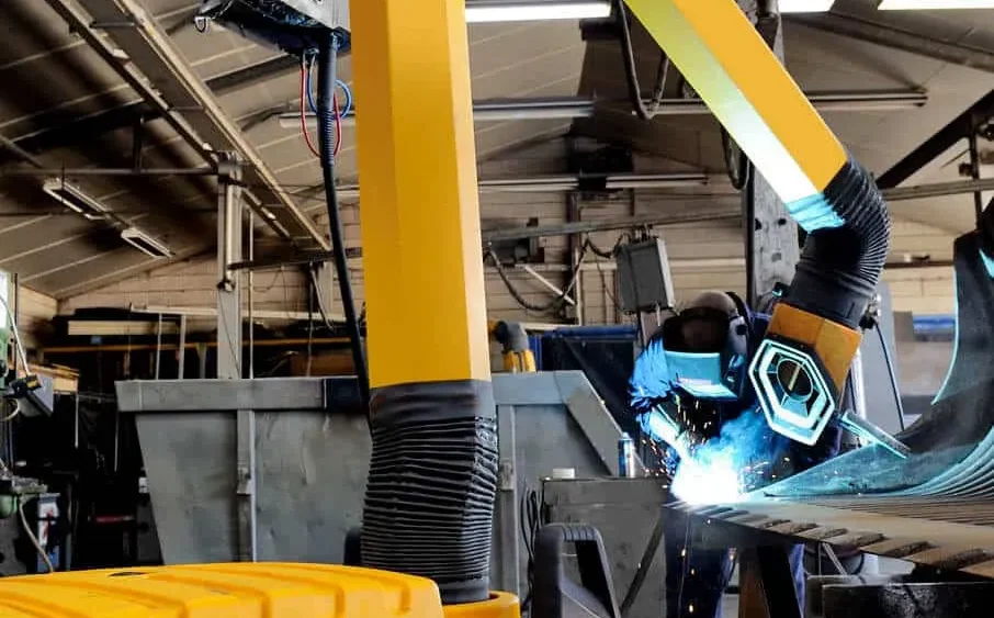 A man welding with a protective mask on a while yellow Remove The Fume fume extractor arm is inhaling all the harmful fume away.