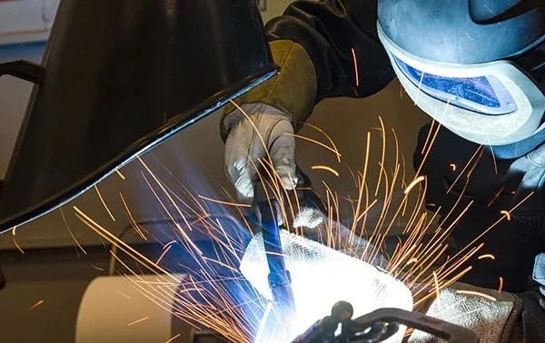 A man welding with a protective mask on and in the background there is a Remove The Fume Fume Extractor who's extraction arm is sucking all the harmful fumes away.