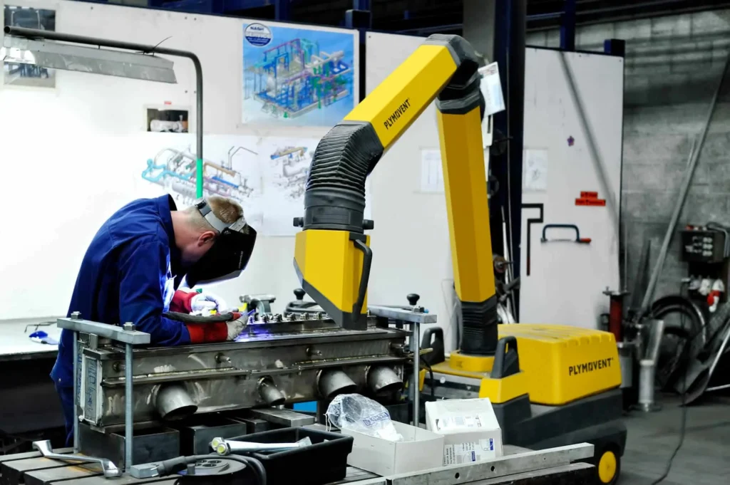 A man welding with a protective mask on a while yellow Remove The Fume fume extractor arm is inhaling all the harmful fume away.