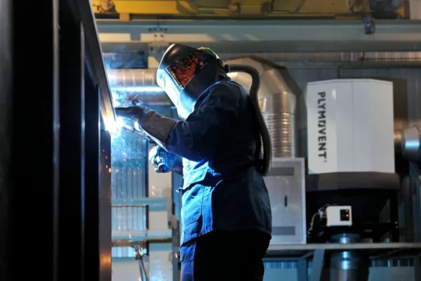 A man welding with a protective mask on while a Plymovent grey colored fume extractor is visible in the background